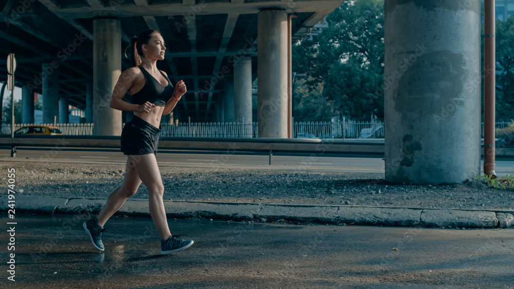 Shot of a Beautiful Fitness Girl in Black Athletic Top and Shorts Jogging in the Street. She is Running in an Urban Environment Under a Bridge with Cars in the Background.