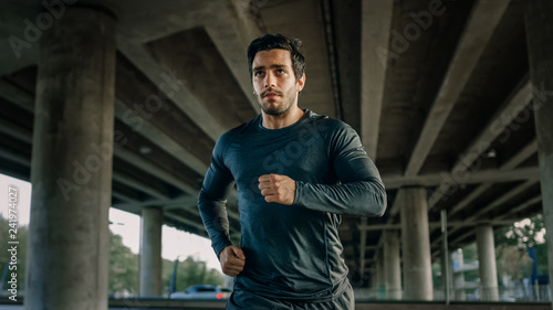 Athletic Young Man in Sports Outfit is Jogging in the Street. He is Running in an Urban Environment Under a bridge with Cars in the Background.