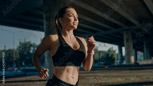 Close Up Shot of a Beautiful Busty Fitness Girl in Black Athletic Top Jogging in a Sunny Street. She is Running in an Urban Environment Under a Bridge with Cars in the Background.