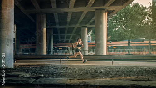 Beautiful Fitness Girl in Black Athletic Top and Shorts is Energetically Running in the Street. She is Running in an Urban Environment Under a Bridge with a Train in the Background.