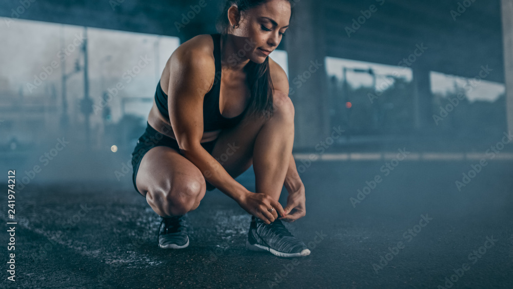 Beautiful Confident Fitness Girl in Black Athletic Top and Shorts Ties Shoelaces. She is in an Urban Environment Under a Bridge with Cars in the Background.