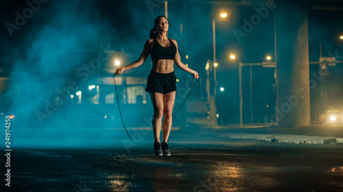 Beautiful Energetic Fitness Girl in Black Athletic Top and Shorts is Skipping/Jumping Rope. She is Doing a Workout in an Evening Foggy Urban Environment Under a Bridge with Cars in the Background. © Gorodenkoff