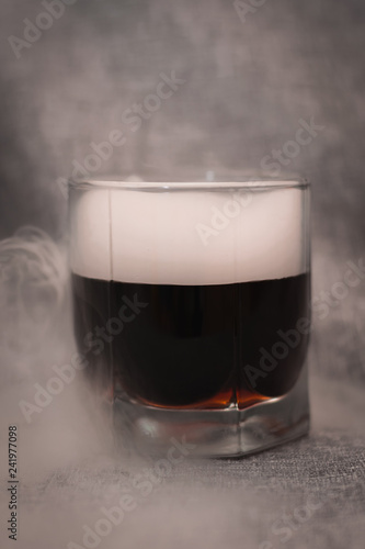 glass of dark liquid and smoke around on a gray background