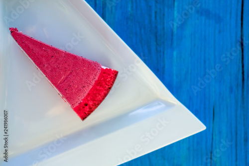 Piece of red berry mousse cake on a white plate in cafe. Sweet dessert on background of blue wooden table, top view.