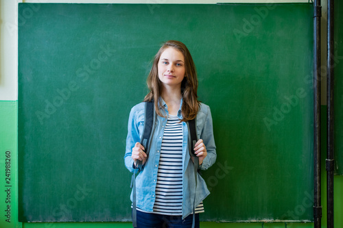 Young confident smiling female high school student standing in front of chalkboard in classroom, wearing backpack, looking at camera. Waist up portrait.