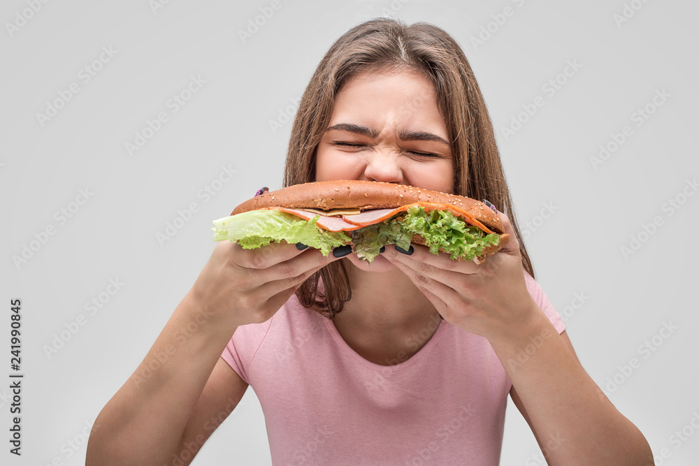 Hungry young woman bite burger. She devour it. Isolated on grey background. Hungry young woman bite burger. She devour it. Isolated on grey background.