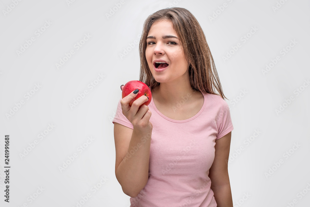 Attractive young woman hold apple in hand. Her mouth is opened. She look straight. Isolated on grey background. Attractive young woman hold apple in hand. Her mouth is opened. She look straight. Isolated on grey background.