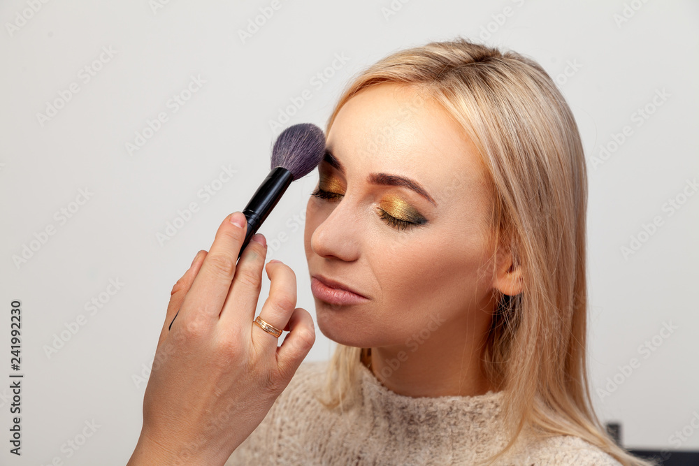 Profile of a blonde model with an oriental-style eye make-up on a white background in the studio, makeup artist with a brush in her hand contours the face with tone, powder and blush. Profile of a blonde model with an oriental-style eye make-up on a white background in the studio, makeup artist with a brush in her hand contours the face with tone, powder and blush.
