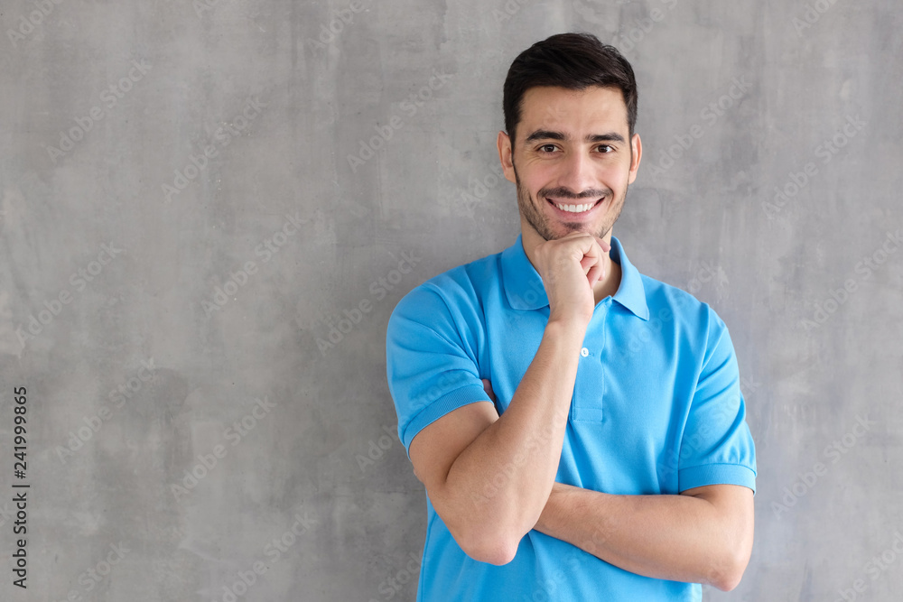 Portrait of smiling handsome man in blue polo shirt, touching his chin, standing against gray textured wall with copy space for your ads Portrait of smiling handsome man in blue polo shirt, touching his chin, standing against gray textured wall with copy space for your ads