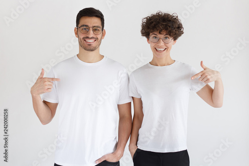 Daylight shot of smiling couple pointing at  blank white t-shirts with index finger, copy space for ads, isolated on gray background