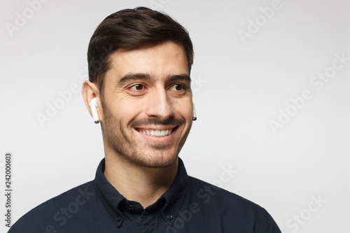 Portrait of smiling young business man listening to music or radio, uses modern wireless earphones, wearing blue shirt. Copy space for text © Damir Khabirov