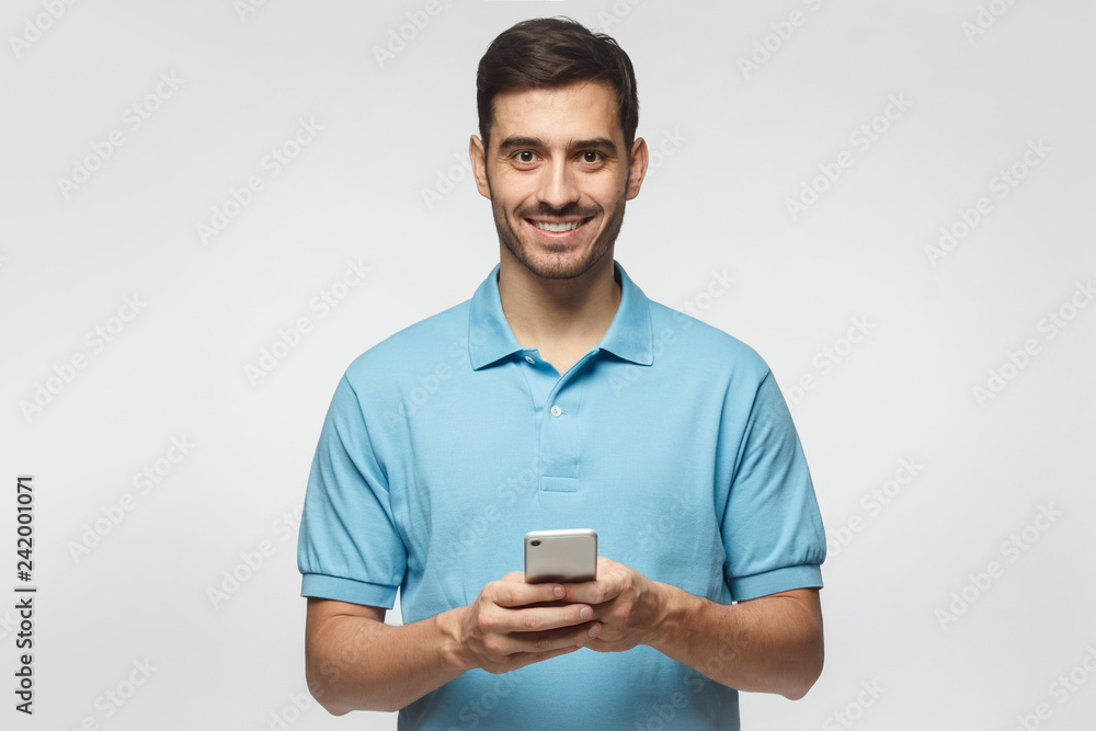Young smiling man standing isolated on grey background, looking at camera, holding phone with both hands Young smiling man standing isolated on grey background, looking at camera, holding phone with both hands