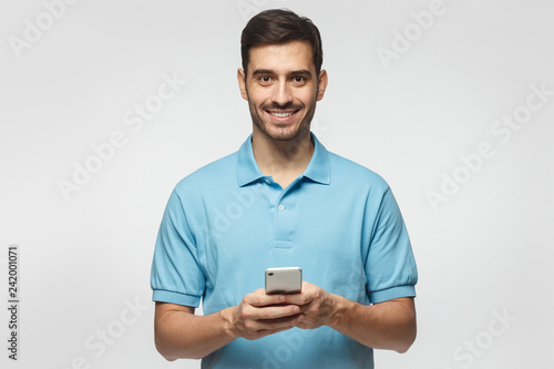 Young smiling man standing isolated on grey background, looking at camera, holding phone with both hands © Damir Khabirov