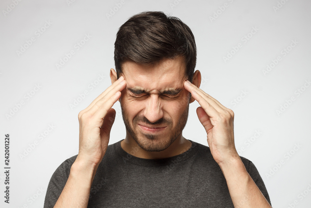 Portrait of young man isolated on gray background, suffering from severe headache, pressing fingers to temples with closed eyes - obrazy, fototapety, plakaty 