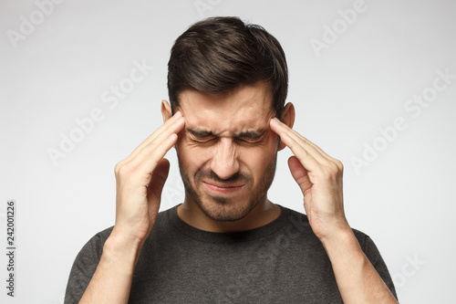 Portrait of young man isolated on gray background, suffering from severe headache, pressing fingers to temples with closed eyes photo