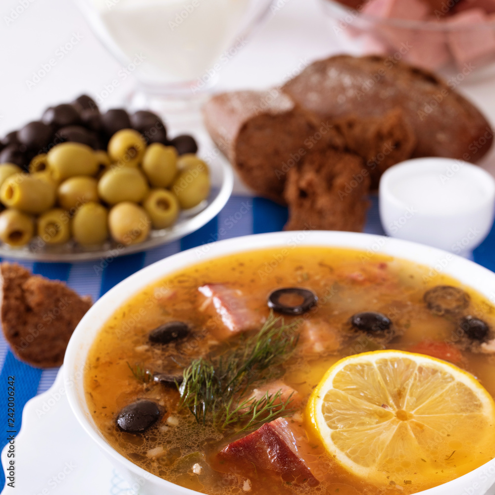 Traditional Russian soup Solyanka cooked with meat, sausages, salted cucumbers on wite bowl. Black bread, lemon, olived and tomatoes on color blue background, homemade food. Traditional Russian soup Solyanka cooked with meat, sausages, salted cucumbers on wite bowl. Black bread, lemon, olived and tomatoes on color blue background, homemade food.