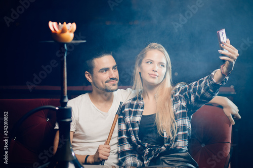 Beautiful couple in nightclub smoking hookah, girl take selfie on smartphone
