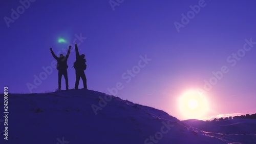 men tourists with backpacks are in winter on hill in rays of bright sun. Climbers on top of snowy mountain rejoice at their successes  raise their hands and happily jump.