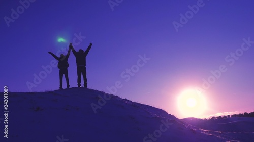 men tourists with backpacks are in winter on hill in rays of bright sun. Climbers on top of snowy mountain rejoice at their successes, raise their hands and happily jump.