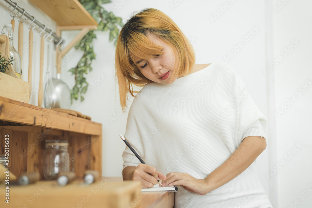 Asian woman write shopping lists in notepad by pen on her kitchen counter at home and reading and tries to remember all ingredients to shopping in grocery. Lifestyle women relax at home concept. Asian woman write shopping lists in notepad by pen on her kitchen counter at home and reading and tries to remember all ingredients to shopping in grocery. Lifestyle women relax at home concept.
