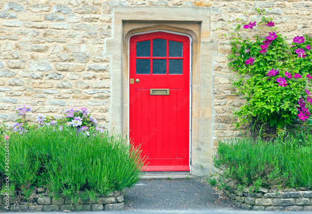 Φωτογραφία Bright red wooden doors in an old traditional English stone house, surrounded by climbing pink clematis on the golden wall, lavender in the front garden