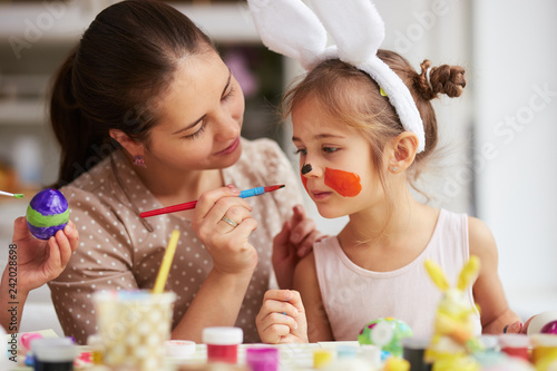 Mother draws on daughter s face while dyeing the eggs for the Easter table in the cozy light kitchen.