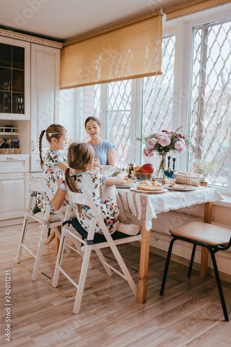 Young caring mother and her two little daughters have a breakfast in the light kitchen with large window