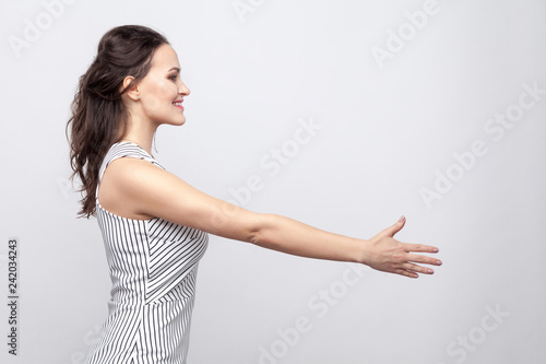 Profile side view portrait of happy beautiful young brunette woman with makeup and striped dress standing and giving hand to greeting and toothy smile. indoor studio shot, isolated on grey background.