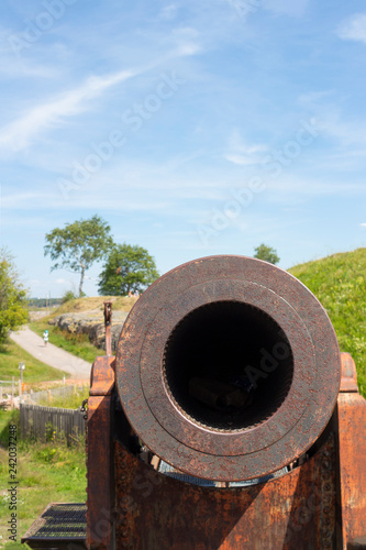 An old cast-iron artillery cannon and granite old fortifications on Suomenlinna fort island are overgrown with grass on a sunny summer day in Finland, and the flag of Finland is on the ancient walls.