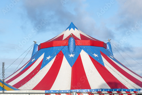 Red and white circus tent topped with bleu starred cover against a sunny blue sky with clouds