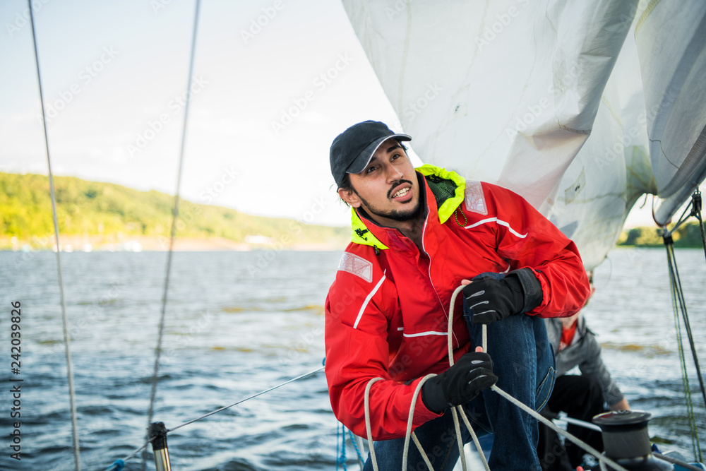 Fototapeta premium Handsome man on yacht touches white sail called assymetric spinnaker, works with tackle in sea at summer day, under view.