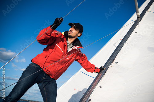 Young man skipper in red windbreaker and black cap sets the sails on a sailing yacht © romankosolapov