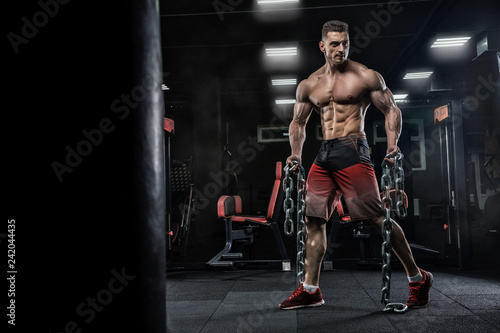 Young handsome male athlete bodybuilder does exercises for leg muscles, uses chains. Beautiful dark background. Concept - gym sports nutrition diet styroyd health simulators strength power crossfit.