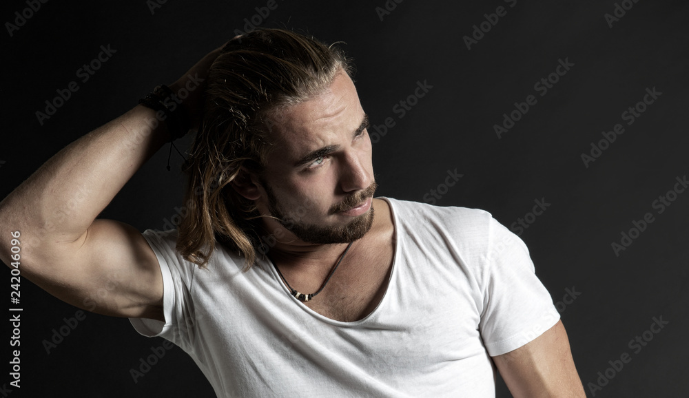 Naklejka premium Portrait von einem jungen Mann mit Bart und langen Haaren in skeptischer nachdenklicher Pose, Studio, dunkler Hintergrund