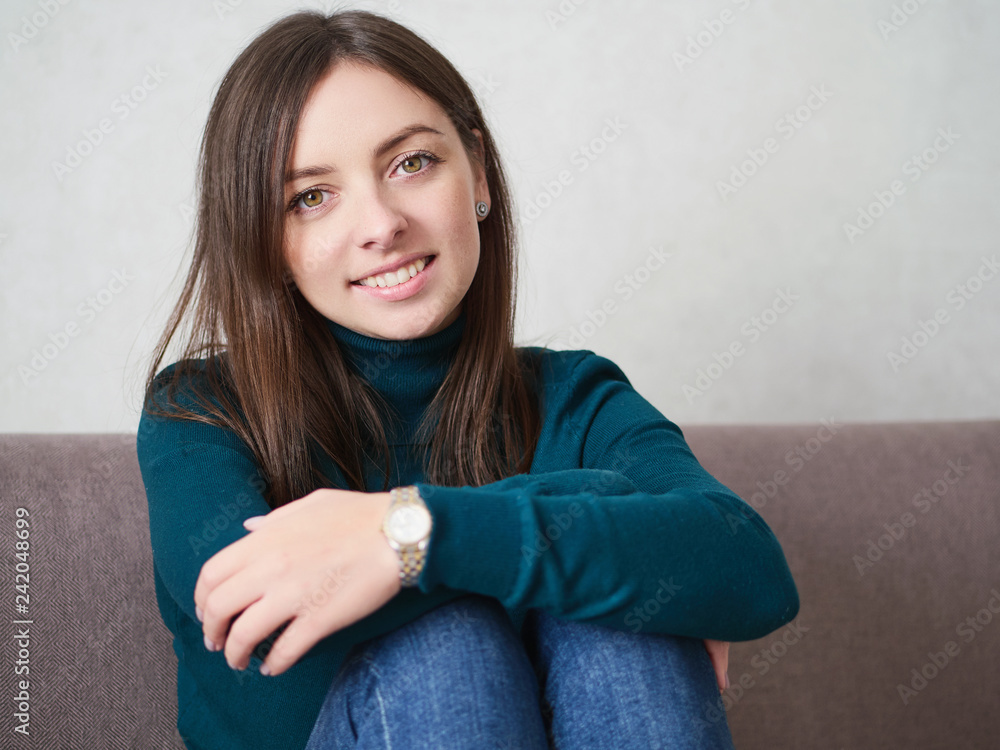 Smiling pretty brunette woman with magnificent eyes and adorable smile in green sweater and blue jeans relaxing sitting on sofa at home Smiling pretty brunette woman with magnificent eyes and adorable smile in green sweater and blue jeans relaxing sitting on sofa at home