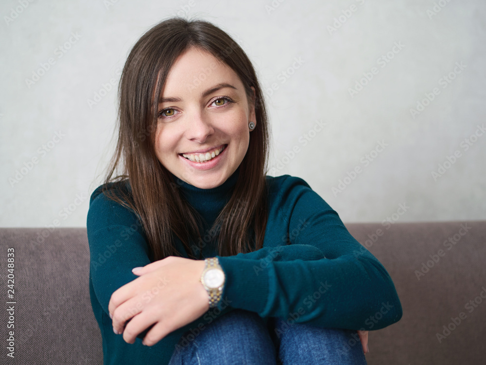 Smiling pretty brunette woman with magnificent eyes and adorable smile in green sweater and blue jeans relaxing sitting on sofa at home Smiling pretty brunette woman with magnificent eyes and adorable smile in green sweater and blue jeans relaxing sitting on sofa at home