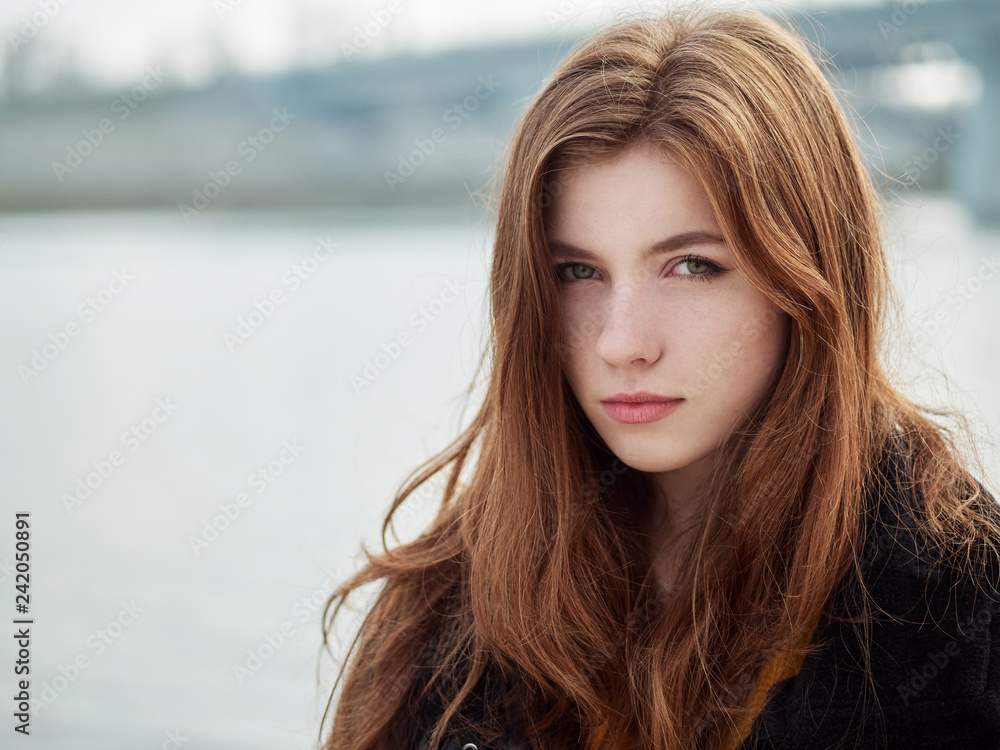 Close up portrait of fabulous redhead woman with long hair in yellow sweater black leather jacket on blurred river pier background.