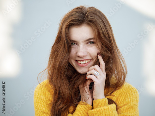 Headshot of fabulous redhead woman with long hair in yellow sweater having fun on grey concrete wall background with sun light spots.