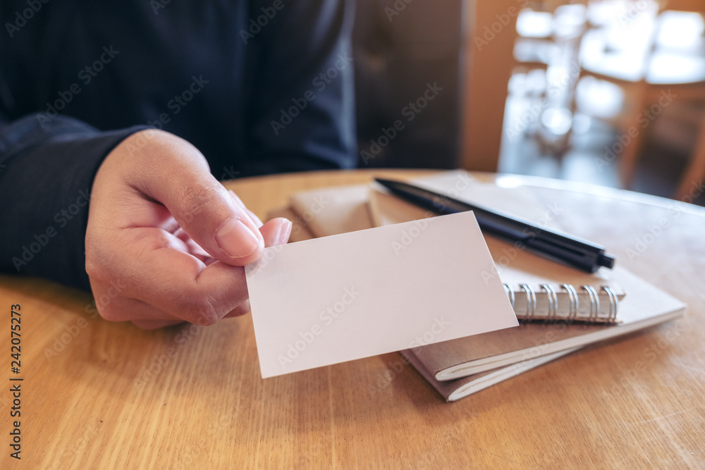 A woman holding and giving a blank empty business card to someone with notebooks on table A woman holding and giving a blank empty business card to someone with notebooks on table