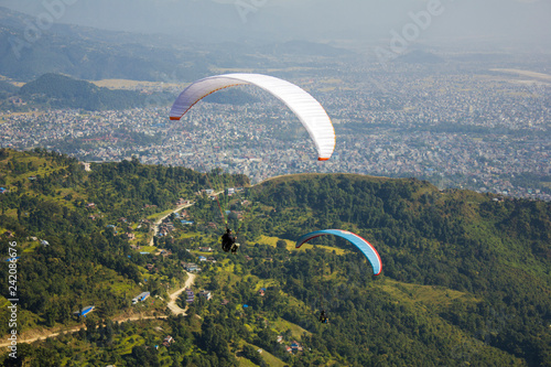 two paragliders on a white and blue parachute fly over the green mountains a city in the valley