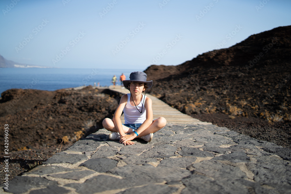 A boy in shorts, a t-shirt and a hat is sitting on a stone path by the sea.
