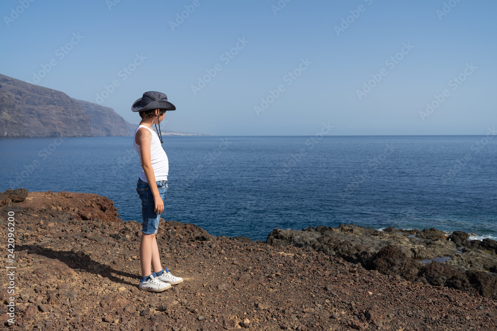 A boy in shorts, a T-shirt and a hat is standing on a rocky seashore and looking into the distance.