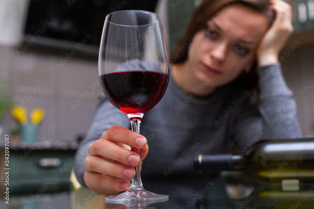 Depressed, divorced crying woman sitting alone in kitchen at home and drinking a glass of red wine because of problems at work and troubles in relationships. Social and life problems