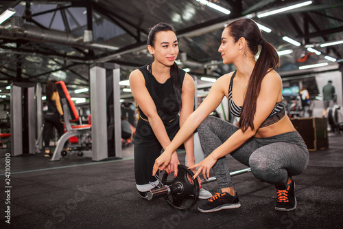 Picture of cheerful young women stand on knees togethr and smile. They look at each other. Yung asian woman help with weight for barbell.