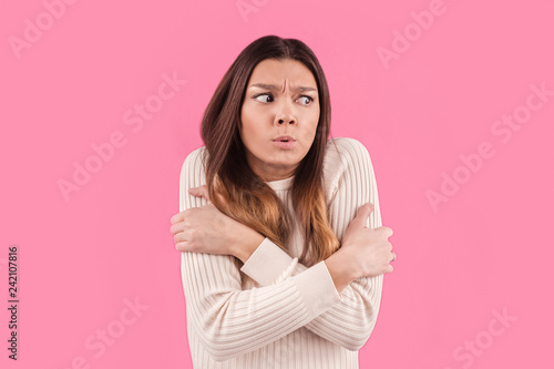 Dissatisfied woman feels cold during freezing chilly weather  hugs herself and clenches teeth  feels discomfort  afraids of something  isolated over pink background. Close up shot