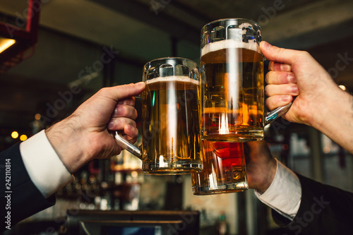 Picture of three mugs of beer in men hands. People wear suits. They are in bar.