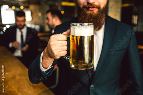 Cut view of bearded man in suit hold beer mug. Other two office workers sit behind.