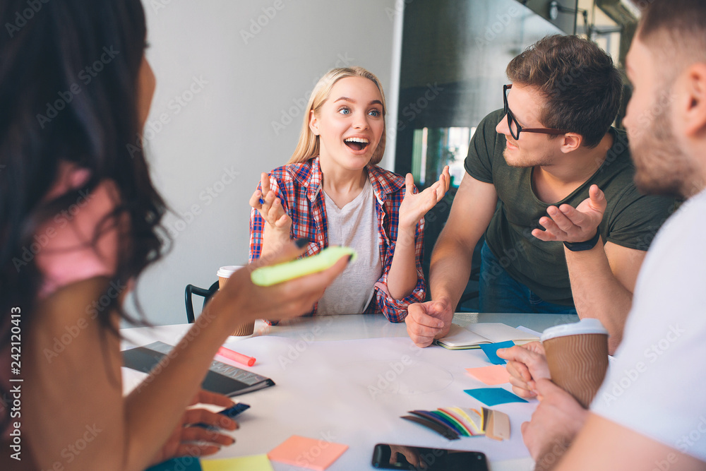 Fototapeta premium Cheerful young blonde woman smile and look forward. She wave with hands. Brunette woman and two young men look at her. They listen to. People work together in coworking.