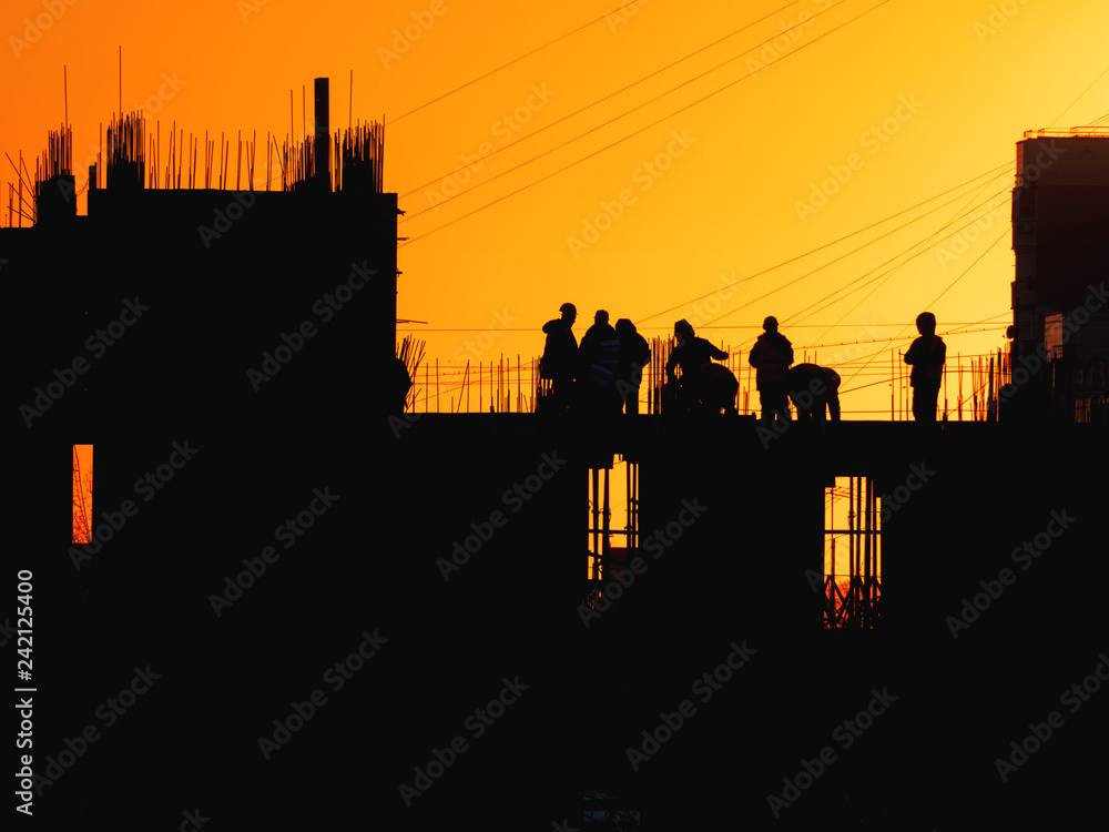 Construction of residential building. Builders go on the unfinished floor with protruding fittings. Silhouettes of workers on the background of an orange sunset.