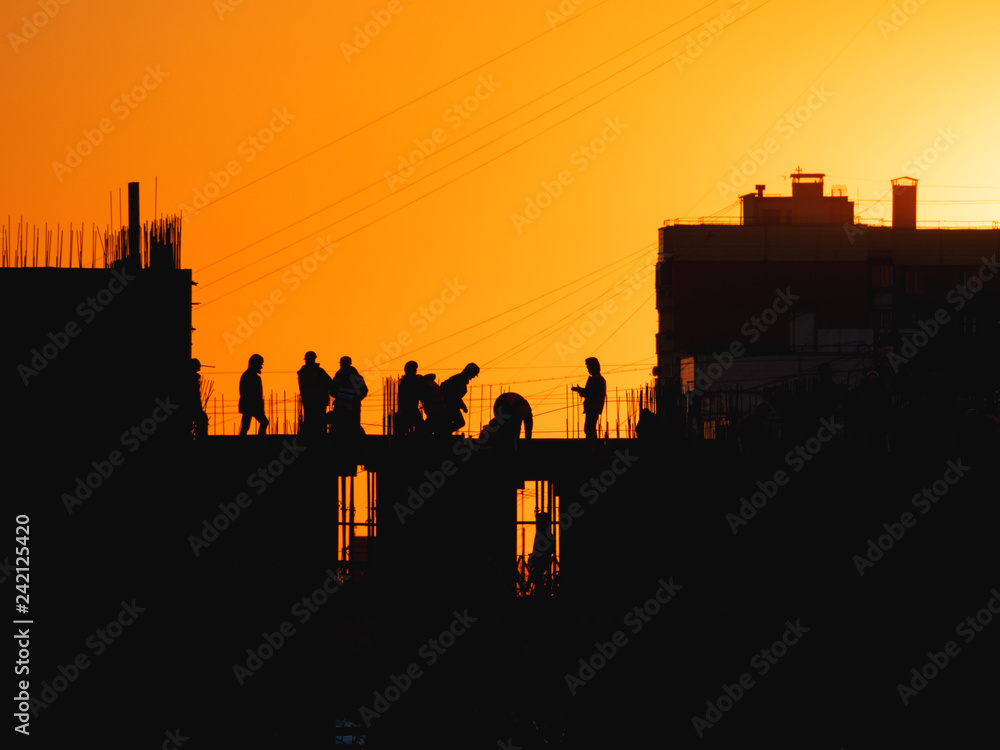 Construction of residential building. Builders go on the unfinished floor with protruding fittings. Silhouettes of workers on the background of an orange sunset. Construction of residential building. Builders go on the unfinished floor with protruding fittings. Silhouettes of workers on the background of an orange sunset.